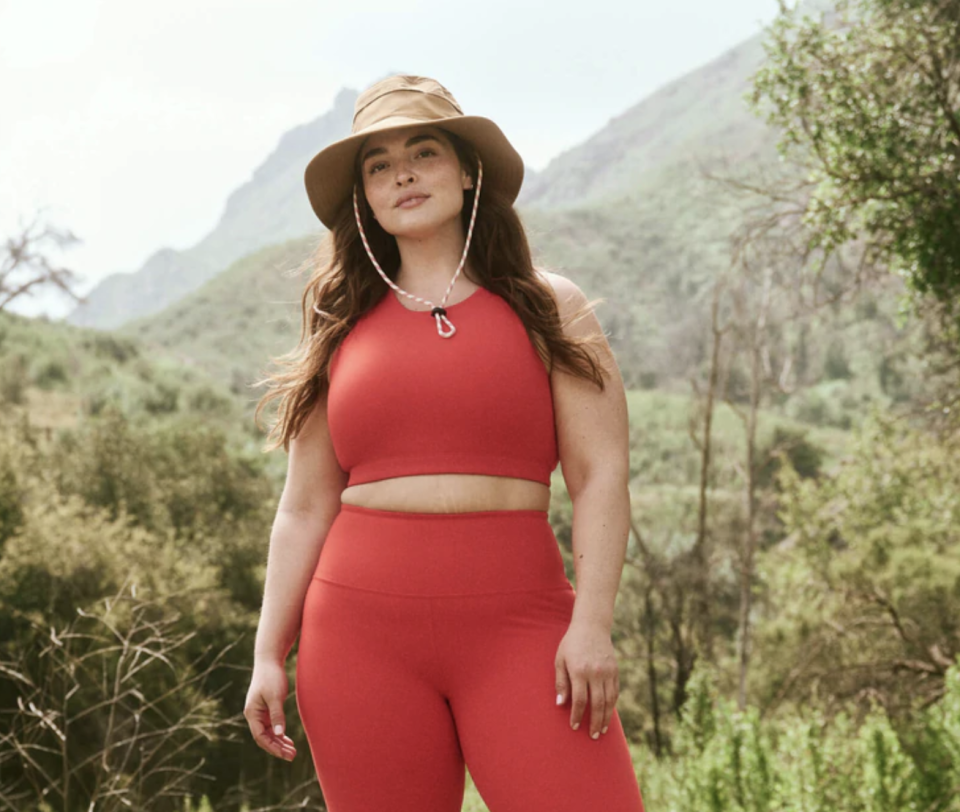 plus size model wearing red top, red leggings and brown hat in mountains and forest, The Great + Eddie Bauer The Hiking Tank in Pimento (Photo via The Great)